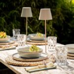 Elegantes Esszimmer-Setup im Freien mit Schüsseln und Tellern auf gewebten Tischsets. Artischocken garnieren die Schüsseln, Kristallwassergläser und beige Servietten mit Lavendelzweigen schmücken den Tisch. Zwei schlanke Akku LED Tischleuchte - Bunte RGB-Lampen werten das Ambiente auf.