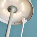 Nahaufnahme des Akku LED Tischleuchtenfußes mit winzigen LEDs. Die Lampe wird über ein USB-Kabel angeschlossen. Sie hat einen Metallfuß und einen hellgrünen Mast vor einem gedämpften blaugrünen Wandhintergrund, was ihr modernes Design unterstreicht.