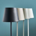 Vier Akku-LED-Tischleuchten mit kegelförmigen Schirmen mit Farbverlauf von Schwarz nach Weiß stehen in einer ordentlichen Reihe vor einem blaugrünen Hintergrund und verfügen über schlanke, gerade Sockel.