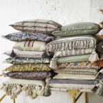 Ein Stapel dekorativer Kissenbezüge – Evie-Kissen mit verschiedenen Mustern und Texturen wie Karo-, Streifen- und Blumenmustern liegt hübsch auf einer hellen Bank. Einige sind mit Quasten und Fransen versehen, was ihren Bohème-Stil vor einer weißen Wand noch verstärkt.