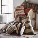 Kissenbezug - Evie-Kissen mit Streifenmuster und Quasten sind auf einem Korbstuhl gestapelt und betonen die Textur des rustikalen Holzbodens. Ein großes Fenster im Hintergrund füllt den Raum mit natürlichem Licht und hebt die erdigen Töne der Kissen hervor.