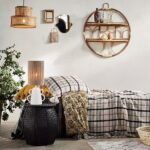 Ein gemütliches Schlafzimmer präsentiert eine karierte Tagesdecke und eine senffarbene Decke mit einem passenden Kissenbezug – Evie. Auf einem schwarzen Beistelltisch stehen eine Lampe und ein Krug. Geflochtene Körbe, Spiegel an der weißen Wand, eine Blattpflanze und runde Regale schaffen eine warme, einladende Atmosphäre.