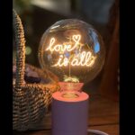 Die Statement-Glühbirne „Love is all“ besteht aus beleuchteten Wörtern auf einem rosa Sockel und einem geflochtenen Korb im Hintergrund, die eine warme, gemütliche Atmosphäre schaffen.