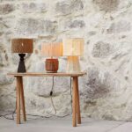 Drei Tischleuchten - Braid-Lampen stehen auf einer Holzkonsole vor einer Steinwand und sind in Erdtönen und mit strukturierter Oberfläche gehalten. Unter der Konsole sind Stromkabel sichtbar.