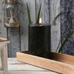 Eine LED Kerze - Authentic - schwarz flackert auf einem Holztablett auf einer rustikalen Oberfläche. Im Hintergrund ist eine verwitterte Wand mit grünen Blättern und einem Glasgefäß mit Muscheln zu sehen.