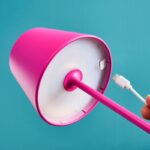Eine Person hebt die rosa Akku LED Tischleuchte - Colorful RGB an und legt den darunter liegenden USB-Ladeanschluss frei. Die Lampe besticht durch ein modernes Design mit leuchtendem Pink auf blaugrünem Hintergrund, während ein USB-Kabel in den Anschluss eingesteckt wird.