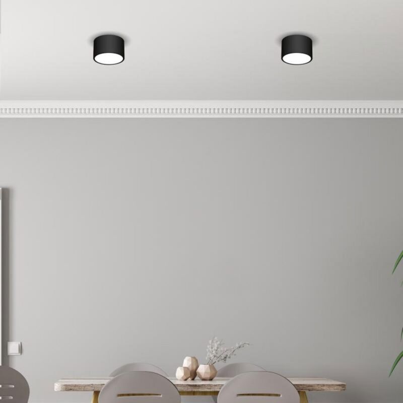 Ein minimalistischer Raum verfügt über eine hellgraue Wand und zwei schwarze Einbauleuchten an der Decke. Ein Holztisch mit drei modernen Stühlen zeigt ein kleines weißes Pflanzenarrangement. Die elegante Aufbauleuchte – Luana wertet das Dekor auf, mit einer grünen Blattpflanze auf der rechten Seite.