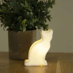 Ein 9 cm großes Akku-LED-Katze-Nachtlicht wirft einen sanften Schein auf eine Holzoberfläche, während eine grüne Topfpflanze in einem runden braunen Behälter der gemütlichen Szene eine natürliche Note verleiht.