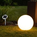 Eine leuchtende LED-Leuchtkugel für drinnen und draußen – Bola SOLAR Sand wirft nachts warmes Licht auf den grasbewachsenen Garten. Ein kleines Solarpanel ragt aus dem hohen Gras hervor, während Korbmöbel den Hintergrund dezent schmücken.