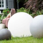 In einer Gartenszene leuchten drei In & Outdoor LED-Leuchtkugeln - Bola SOLAR Sand in verschiedenen Größen auf dem Gras, während im Hintergrund ein Stuhl mit einer rosa Decke und einer weißen Korbtasche zwischen üppigem Grün eingebettet ist.