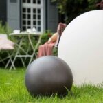 In einer Gartenumgebung stehen im Hintergrund ein kleiner runder Tisch und ein Stuhl mit einer rosa Decke. Im Vordergrund beleuchten eine dunkelgraue Kugel und die In & Outdoor LED Leuchtkugel Bola SOLAR sand sanft das Gras.