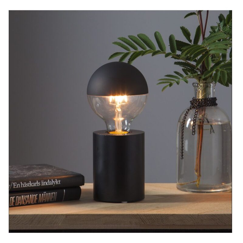 Eine moderne schwarze Leuchtenfuß-Mio-Tischlampe mit runder Glühbirne steht auf einer Holzoberfläche neben gestapelten Büchern. Daneben steht eine Glasvase mit grünem Blattwerk, die mit sanfter Beleuchtung eine minimalistische Szene erzeugt.