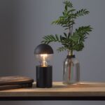 Eine kleine schwarze Lampe mit runder Glühbirne und elegantem Leuchtenfuß – Mio-Sockel steht neben einer Glasvase mit grünen Zweigen auf einem Holztisch, begleitet von einem geschlossenen Buch. Die Szene steht vor einer schlichten grauen Wand.