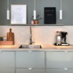 Eine moderne Küche verfügt über hellgrüne Schränke, eine Edelstahlspüle und eine sechseckige Fliesenrückwand. Darüber befindet sich ein Regal mit Dekor und einem Kaffeeschild. Auf der Theke steht eine Kaffeemaschine neben Schneidebrettern und einer rosa Aufbewahrungsbox. Drei MIB 6 Pendelleuchten hängen elegant darüber.
