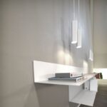 Ein minimalistisches schwebendes Regal mit Büchern und Deko ist an einer hellgrauen Wand montiert. Darüber hängen drei zylindrische Pendelleuchten – MIB 6 in einer Reihe und spenden sanftes Licht. Der Raum strahlt eine moderne und schlichte Designästhetik aus.