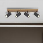 Der Deckenstrahler – Dari 4er verfügt über einen rechteckigen Holzsockel und vier verstellbare schwarze zylindrische Lichter, die an einer weißen Decke über einer dunkelbraunen Wand montiert sind.