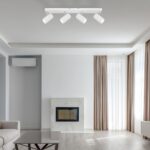 Ein modernes Wohnzimmer ist mit einem Dari 4er Deckenstrahler, beigen Vorhängen und einem grauen Sofa ausgestattet. Hellgraue Wände ergänzen die wandmontierte Klimaanlage über dem weißen Marmorkamin, während große Fenster den Raum mit natürlichem Licht durchfluten und so für eine einladende Atmosphäre sorgen.