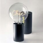 Eine moderne Lampe mit einem schwarzen zylindrischen AKKU Leuchtenfuß zeigt eine große, klare Glühbirne mit „Chic“ im Inneren. Ein zweiter schwarzer zylindrischer Behälter ergänzt das schlichte Setup auf einer weißen Oberfläche und hebt die Statement Bulbs-Kollektion hervor.