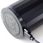 Eine Nahaufnahme eines zylindrischen schwarzen Lautsprechers mit einem seitlich eingesteckten Ladekabel unterstreicht das exquisite Design. Die Unterseite weist eine silberne runde Platte mit eingravierten Spezifikationen auf, die an die robuste Technik der AKKU Leuchtenfuß - Statement Bulbs erinnert.