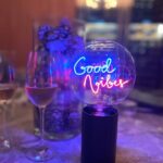 Eine schwach beleuchtete Szene zeigt das Schild „Statement Bulb - Good Vibes“ in einem durchsichtigen, runden Behälter auf schwarzem Sockel. In der Nähe verstärken zwei Weingläser und blassviolette Blumen die Atmosphäre.