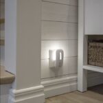 Ein kleines LED-Nachtlicht mit Bewegungs-/Dämmungssensor ist in eine Steckdose an einer weiß getäfelten Wand in der Nähe einer Treppe eingesteckt und strahlt ein sanftes Licht aus, das den Bereich erhellt. Auf einem Regal rechts steht ein geflochtener Korb.