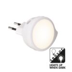 Das LED-Nachtlicht mit Bewegungs-/Dämmerungssensor, Steckdose 2700K ist ein rundes Nachtlicht mit weiß leuchtender Oberfläche. Es verfügt über ein Sensorsymbol und den Text „Leuchtet bei Dunkelheit“ unten rechts, was darauf hinweist, dass es bei schlechten Lichtverhältnissen automatisch leuchtet.