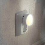 Das LED-Nachtlicht mit Bewegungs-/Dämmerungssensor, Steckdose 2700K wird in eine weiße Steckdose an einer strukturierten beigen Wand eingesteckt. Es strahlt ein weiches, warmes Licht aus, das den Bereich sanft beleuchtet, während sein Design den subtilen Charme der Wand unterstreicht.