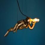 Die Deckenleuchte „Pendelleuchte – Dive-Monkey“ zeigt einen goldenen Taucher mit Taucherbrille und Atemgerät, der anmutig vor einem dunkelblaugrünen Hintergrund eine brennende Glühbirne hält.