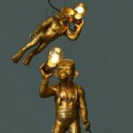 Die Pendelleuchte - Dive-Monkey besteht aus zwei goldenen Lampen in Affenform: eine in Tauchpose, die eine leuchtende Glühbirne hält, als würde sie springen, und die andere steht aufrecht. Beide sind vor einem dunklen Hintergrund angebracht, wodurch ein auffälliger visueller Kontrast entsteht.