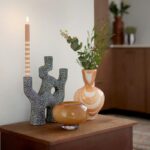 Auf einem Holztisch steht eine brennende Kerze im Kerzenhalter - Trio, eine runde Vase aus bernsteinfarbenem Glas und eine hohe gestreifte Vase mit Grünpflanzen. Im Hintergrund sind ein Holzschrank und verschwommenes Dekor zu sehen.