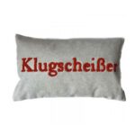 Der Kissenbezug - Klugscheißer ist ein rechteckiger grauer Kissenbezug mit dem elegant in Rot gestickten Wort „Klugscheißer“ in der Mitte.