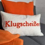 Der Kissenbezug - Klugscheißer, ein beiges Kissen mit dem roten Schriftzug „Klugscheißer“, liegt auf einem grauen Sofa. Ein orangefarbenes Kissen und eine Decke dahinter verleihen der Umgebung lebendige Akzente.