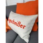 Eine Nahaufnahme des Kissenbezugs – Klugscheißer mit dem Schriftzug „Klugscheißer“ in roter Schreibschrift liegt auf einem grauen Sofa, wobei leuchtend orangefarbene Kissen dem Hintergrund Farbe verleihen.