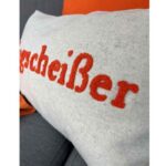 Vor einem grau-orangefarbenen Hintergrund bringt ein weißes Kissen mit dem in kräftigem Rot gestickten Text „Klugcheißer“ eine launische Note in die Szene.
