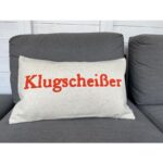 Ein grauer Kissenbezug – Klugscheißer liegt auf einem dunkelgrauen Sofa und zeigt das Wort „Klugscheißer“ in kräftigen roten Buchstaben.