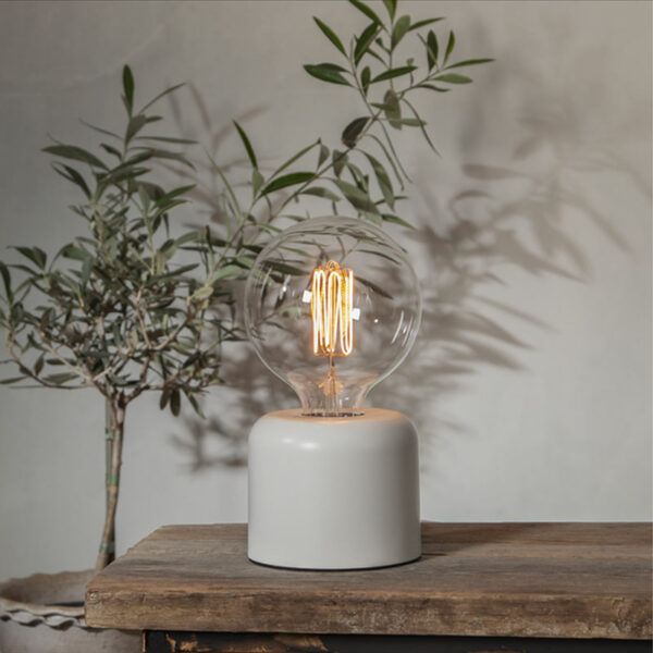 Ein Leuchtmittel im Vintage-Stil - Globe 3,8 W 1800 K dimmbar Ø12,5 cm mit sichtbarem Glühfaden ruht auf einem modernen zylindrischen weißen Sockel auf einem rustikalen Holztisch, während eine kleine Blattpflanze im Hintergrund für Grün sorgt.