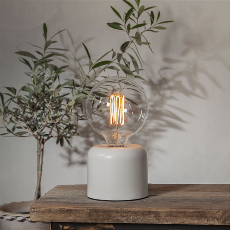 Ein Leuchtmittel im Vintage-Stil - Globe 3,8 W 1800 K dimmbar Ø12,5 cm mit sichtbarem Glühfaden ruht auf einem modernen zylindrischen weißen Sockel auf einem rustikalen Holztisch, während eine kleine Blattpflanze im Hintergrund für Grün sorgt. Ein Leuchtmittel im Vintage-Stil - Globe 3,8 W 1800 K dimmbar Ø12,5 cm mit sichtbarem Glühfaden ruht auf einem modernen zylindrischen weißen Sockel auf einem rustikalen Holztisch, während eine kleine Blattpflanze im Hintergrund für Grün sorgt.