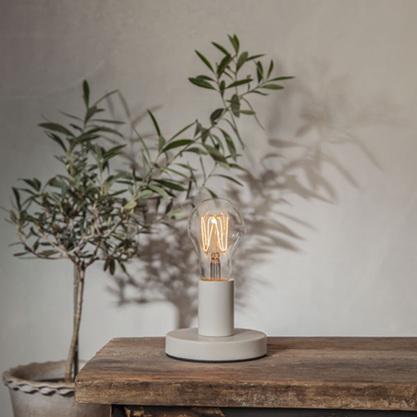 Eine dimmbare 3,8W 1800K Leuchtmittel-Tropfen-Glühbirne mit Ø6cm Durchmesser auf einem minimalistischen runden Lampensockel sitzt elegant auf einem Holztisch. Eine Topfpflanze im Hintergrund wirft ihren Schatten auf die schlichte Wand und verstärkt die gemütliche und rustikale Atmosphäre.
