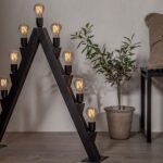 Eine moderne dreieckige Bogen-Stehlampe mit Leuchtmittel – Tropfen 3,8 W 1800 K dimmbar Ø6 cm an jeder Seite steht auf einem Holzboden neben einer Topfpflanze und einem gepolsterten Stuhl vor einer beigen Wand.
