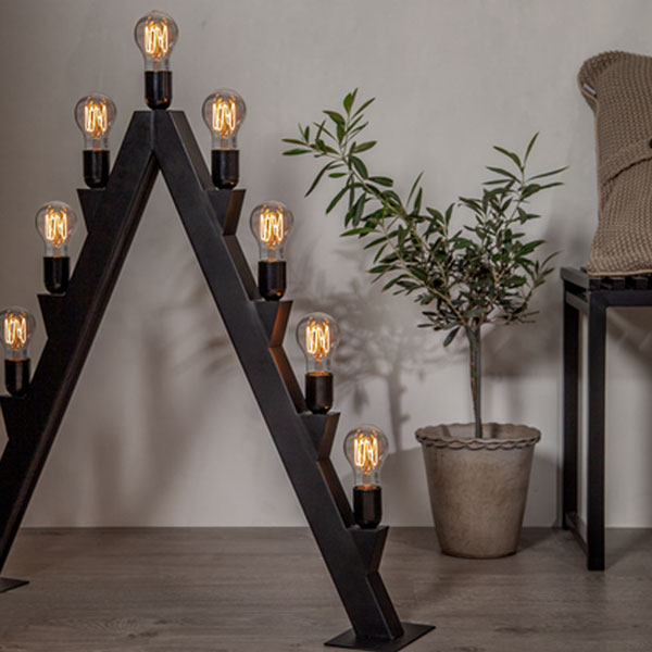 Eine moderne dreieckige Bogen-Stehlampe mit Leuchtmittel – Tropfen 3,8 W 1800 K dimmbar Ø6 cm an jeder Seite steht auf einem Holzboden neben einer Topfpflanze und einem gepolsterten Stuhl vor einer beigen Wand.