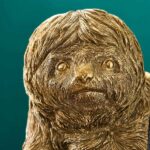 Eine goldene Skulptur eines Faultiers namens Fauli mit detaillierter Fellstruktur, großen Augen und ernstem Gesichtsausdruck hängt wie eine Pendelleuchte vor einem soliden blaugrünen Hintergrund, was seinen metallischen Glanz noch verstärkt.