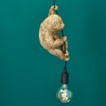 Die Pendelleuchte - Fauli besteht aus einer goldenen Faultierfigur, die an einer schwarzen Kordel hängt und an der eine klare Glühbirne hängt, die vor einem türkisfarbenen Hintergrund ein warmes Licht ausstrahlt.