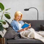 Eine blonde Frau liest ein blaues Buch auf einem grauen Sofa, eingehüllt in eine Decke. Der Raum wird von der modernen schwarzen Stehleuchte - Aike beleuchtet und eine große grüne Pflanze sorgt für Frische. Die weiße Backsteinwand verstärkt die gemütliche Atmosphäre.