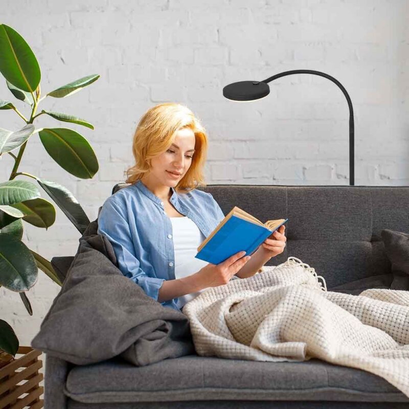 Eine blonde Frau liest ein blaues Buch auf einem grauen Sofa, eingehüllt in eine Decke. Der Raum wird von der modernen schwarzen Stehleuchte - Aike beleuchtet und eine große grüne Pflanze sorgt für Frische. Die weiße Backsteinwand verstärkt die gemütliche Atmosphäre.