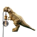Die Tischleuchte - T-Rex, eine goldene Lampe in Dinosaurierform, trägt vor einem schlichten weißen Hintergrund eine leuchtende Glühbirne in ihrem Maul, die über ein elegantes schwarzes Kabel mit der Glühbirne verbunden ist.