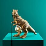 Die Tischleuchte – T-Rex ist eine goldene Dinosaurierfigur, die auf einem blaugrünen Sockel vor einem passenden Hintergrund eine Glühbirne im Maul hält, wobei die Glühbirne durch ein elegantes schwarzes Kabel verbunden ist.