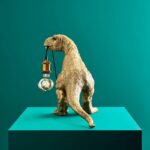 Eine goldene Tischleuchte in T-Rex-Form steht auf einem blaugrünen Sockel und hält eine Glühbirne mit freiliegendem Glühfaden. Der einfarbig blaugrüne Hintergrund verstärkt die verspielte und moderne Ausstrahlung.