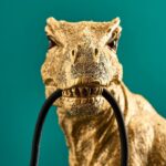 Die Tischleuchte - T-Rex besticht durch ein goldenes Dinosaurierdesign, das vor einem blaugrünen Hintergrund ein schwarzes Kabel im Maul hält, wobei seine strukturierte Oberfläche und die lebendige Farbe einen starken Kontrast zum dunklen Kabel bilden.