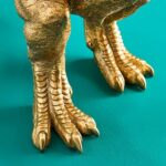 Nahaufnahme goldener T-Rex-ähnlicher Füße mit scharfen Krallen, die an einen einzigartigen Tischleuchte-T-Rex-Sockel erinnern, vor einem türkisfarbenen Hintergrund. Die detaillierten Texturen imitieren schuppige Haut.