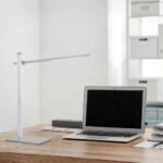 Ein moderner Arbeitsplatz mit einer eleganten, silbernen Tischleuchte - Leve und einem offenen Laptop auf einem Holzschreibtisch. Im Hintergrund sind weiße Regale mit ordentlich angeordneten Kisten zu sehen.